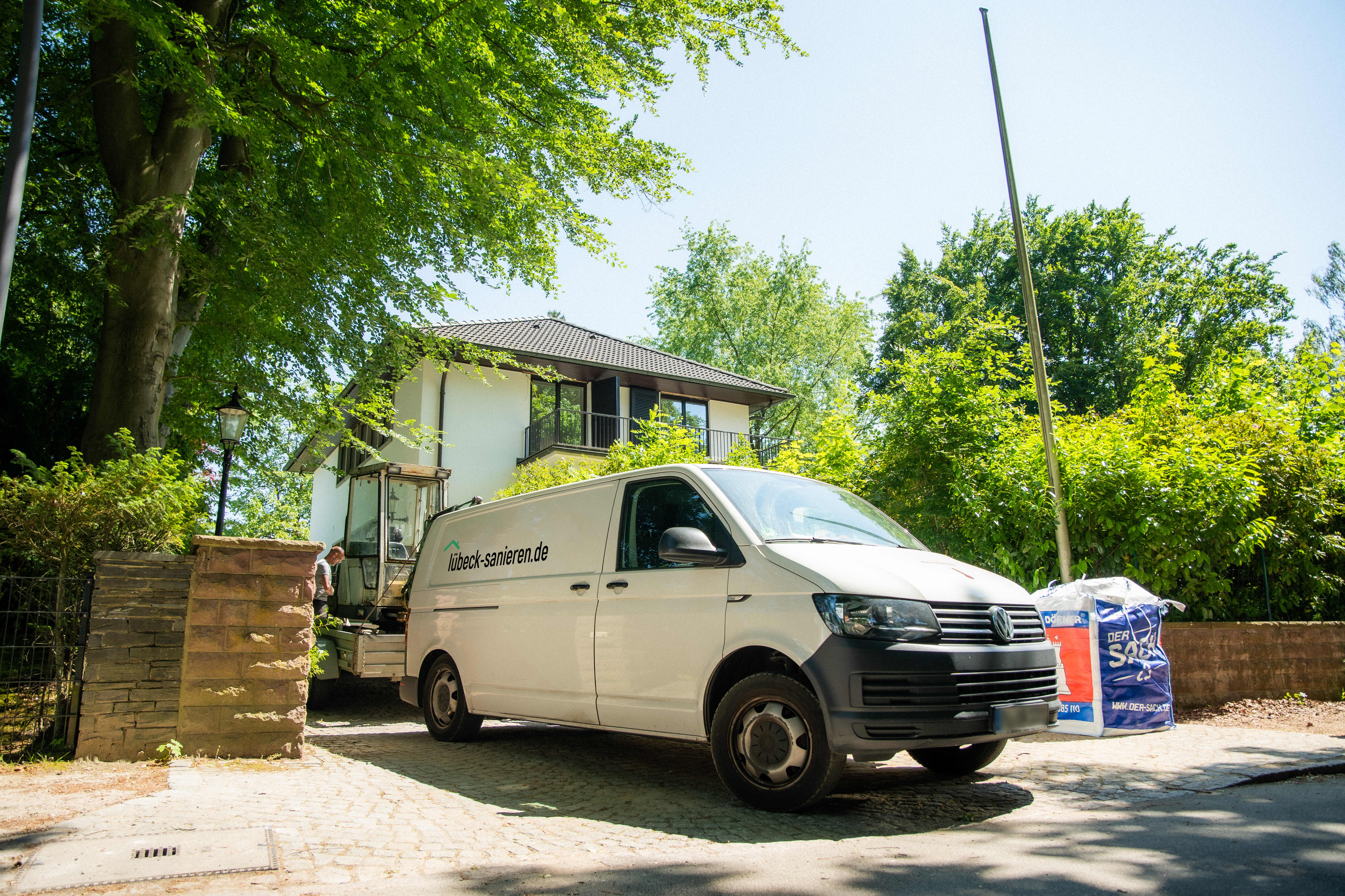 Transporter von luebeck-sanieren.de vor einem Haus, bereit für Sanierungsarbeiten, umgeben von Bäumen und Baustellenmaterial.