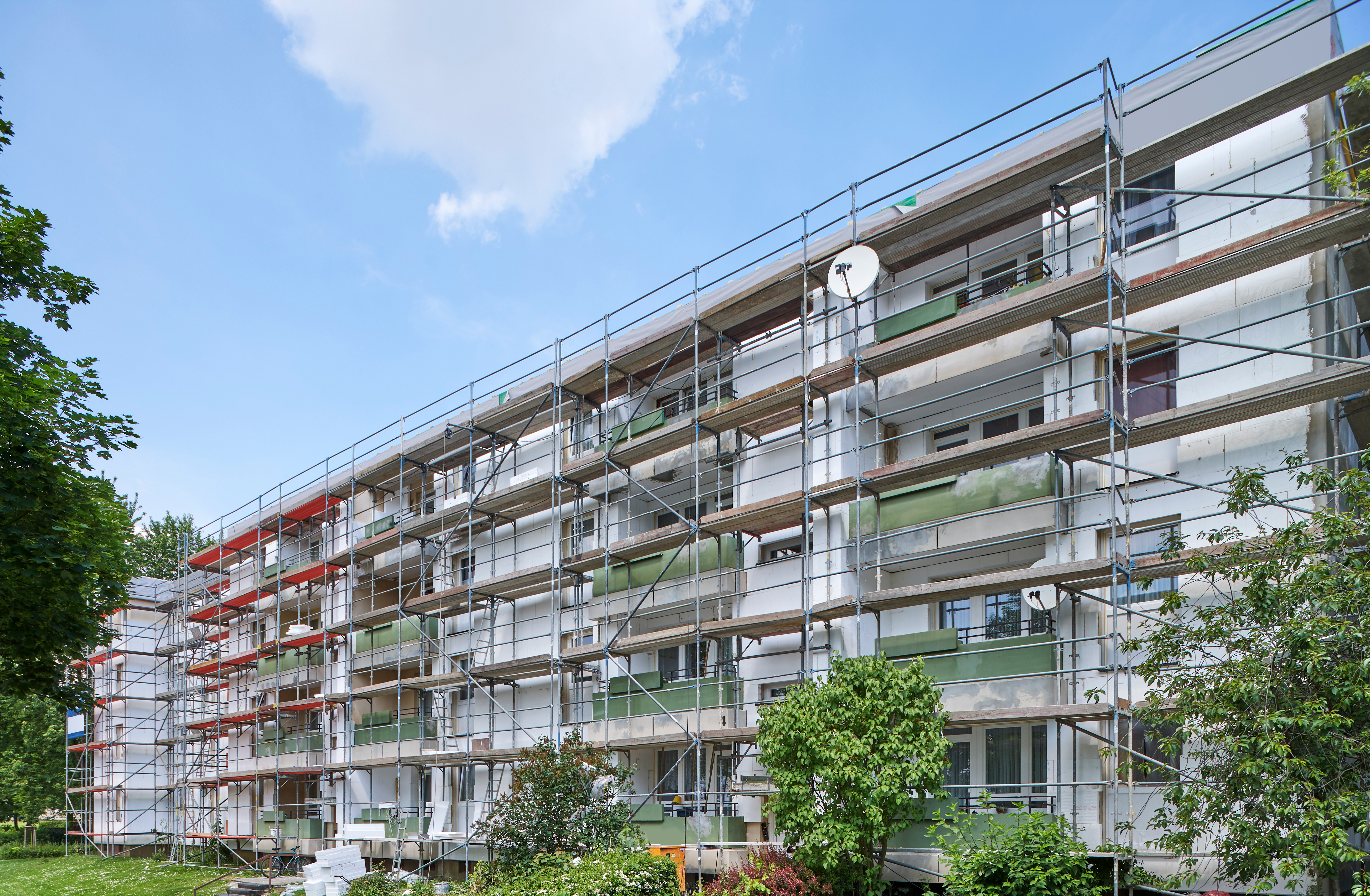 Mehrfamilienhaus während einer Fassadensanierung, eingerüstet und bereit für Renovierungsarbeiten, mit grünem Umfeld und blauem Himmel.